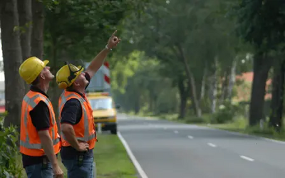 2 mensen met veiligheidshesjes aan de kant van de weg, 1 man wijst naar boven